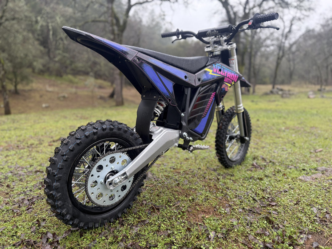 An electric dirt bike with black and blue plastics, labeled 'Voltron Edition', featuring large off-road tires, a visible brushless motor, and a digital display.