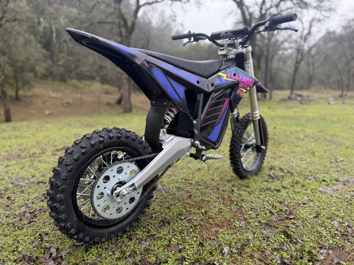 An electric dirt bike with black and blue plastics, labeled 'Voltron Edition', featuring large off-road tires, a visible brushless motor, and a digital display.