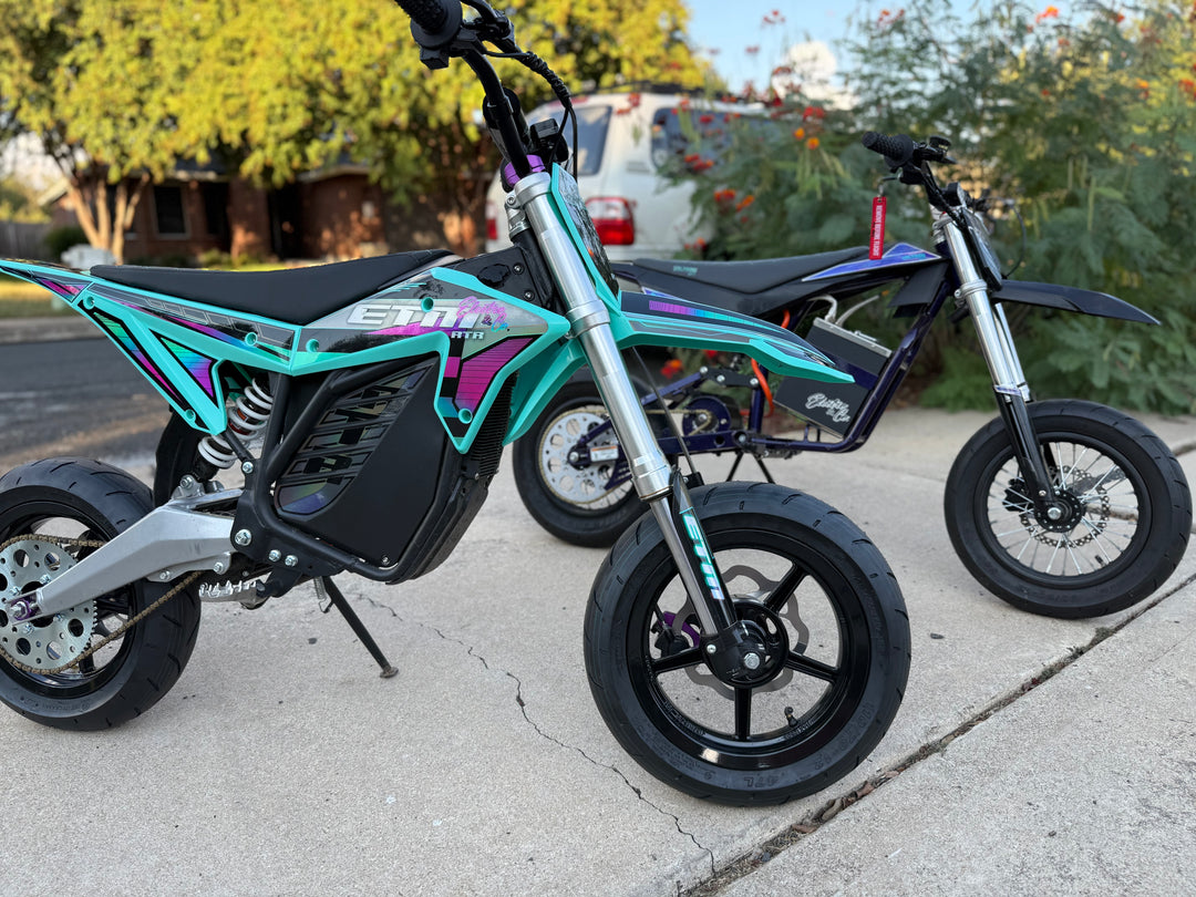 Two small off-road motorcycles on a sidewalk with a residential background.