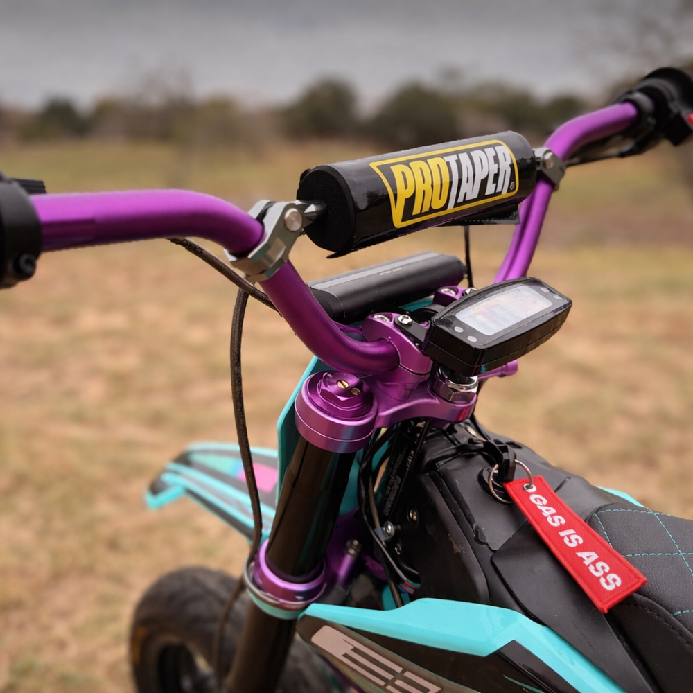 Close-up of a dirt bike handlebar with ProTaper suspension and 'Gas is Ass' tag, set against a blurred natural background.