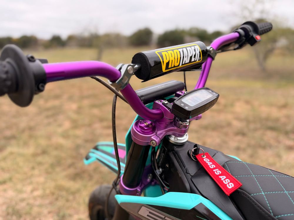 Close-up of a motorcycle handlebar with ProTaper and Yous & Ass branding, set against a blurred outdoor background.