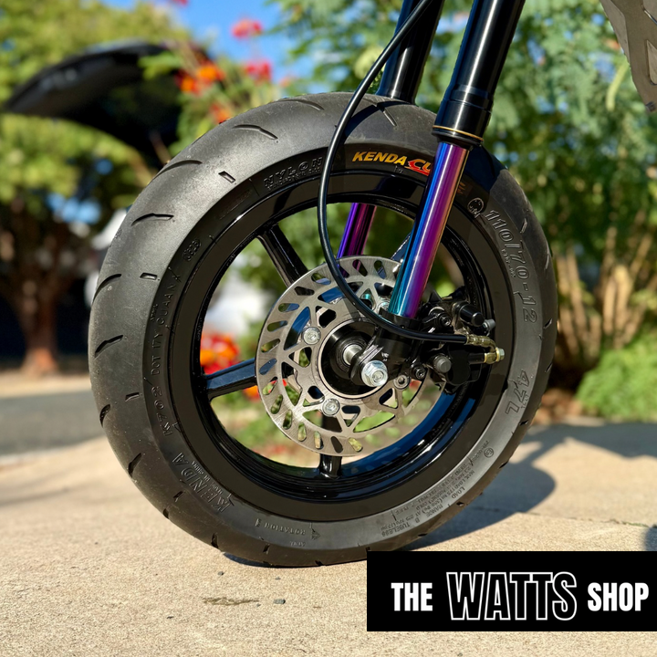 Close-up of a motorcycle wheel with Kenda tire and disc brake, blurred background with 'The Watts Shop' branding.