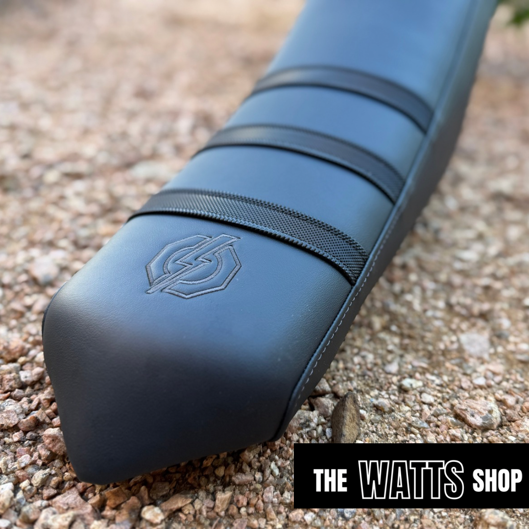 Blue motorcycle seat with a logo on a gravel surface, branded 'The Watts Shop'.