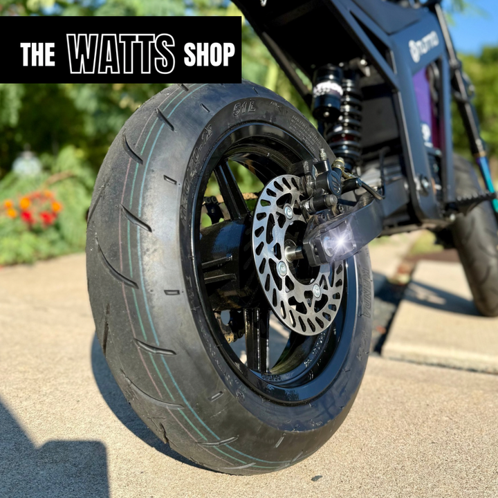 Close-up of a motorcycle wheel with 'The Watts Shop' branding in the background.